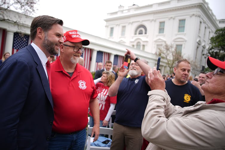 JD Vance, in a suit and tie, poses for a photo with a man in a United Auto Workers t-shirt.