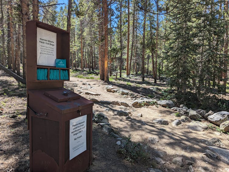 A brown box stands near a trail in the forest. Numerous turquoise bags are folded and placed on shelves. A sign, with black lettering on white laminated paper, is attached to the kiosk. One reads: