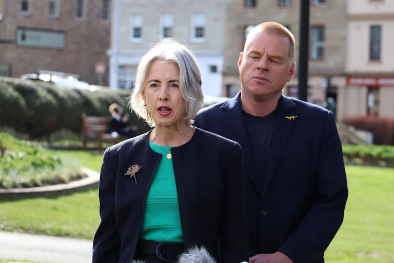 Two politicians, one female and one male, stand in front of microphones and speak to the media