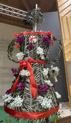 A decorated woven symbol of Persephone made of leaves, flowers and red ribbon.