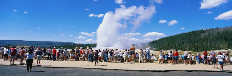 A large group of people stand in a line looking at an area where steam is rising from the ground.