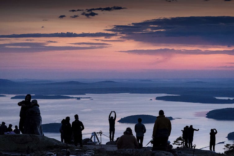 A group of people stand on a mountaintop looking at a bay with islands as the sun rises over the horizon.
