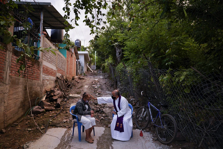 A seated man in a white robe reaches over to touch the forehead of a seated woman as they sit in an alleyway outside.