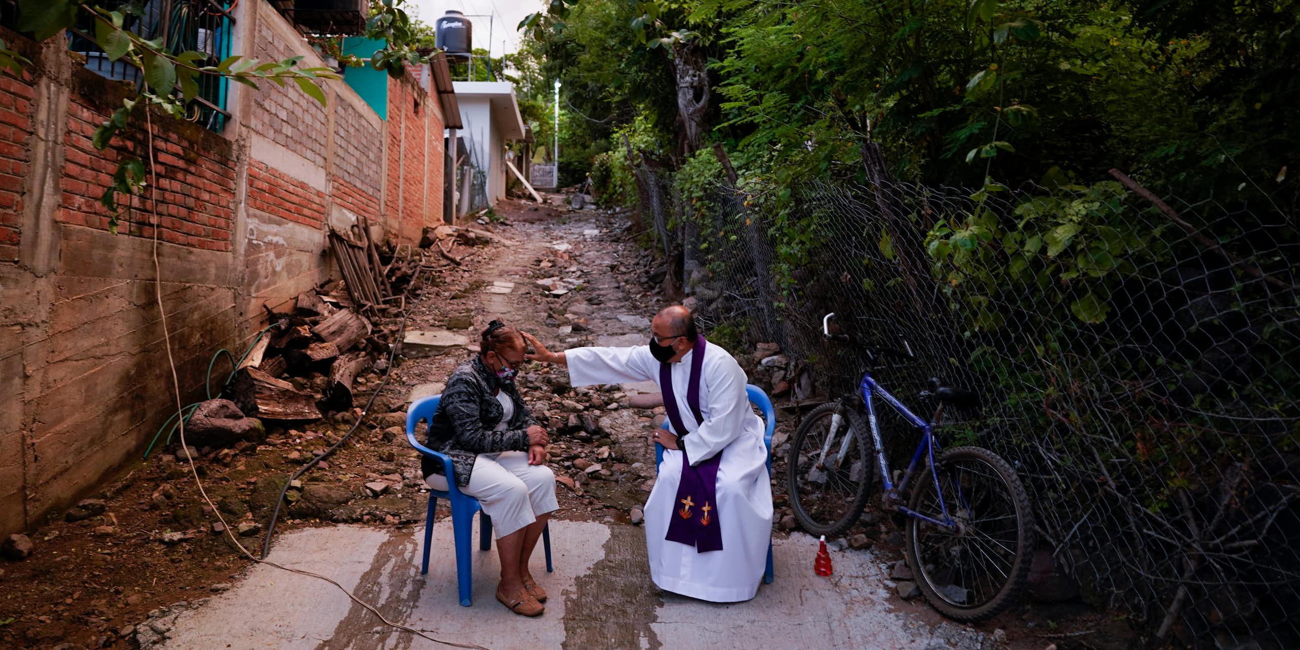 A seated man in a white robe reaches over to touch the forehead of a seated woman as they sit in an alleyway outside.