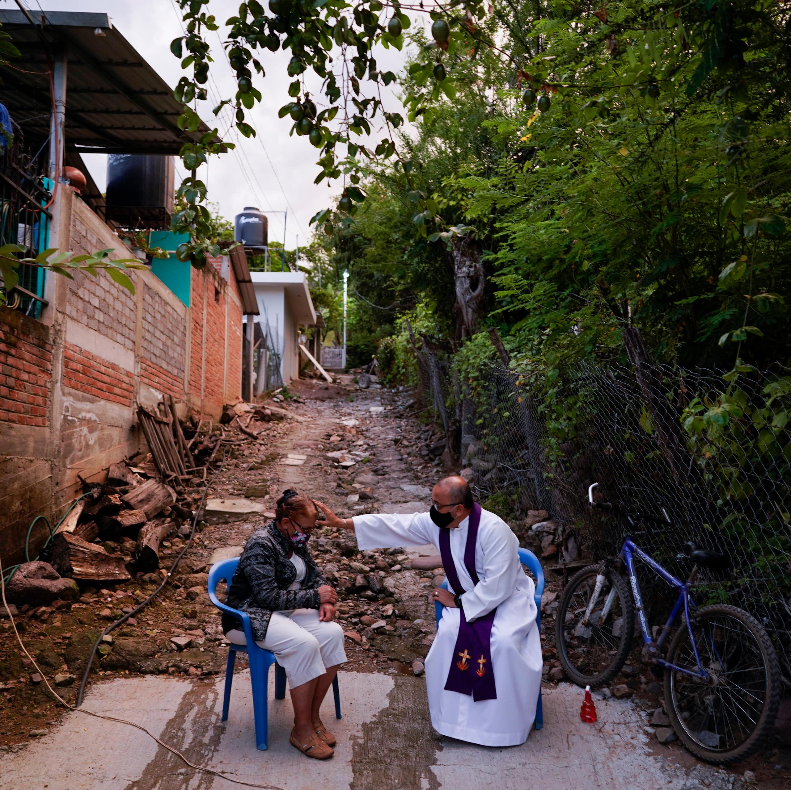 A seated man in a white robe reaches over to touch the forehead of a seated woman as they sit in an alleyway outside.