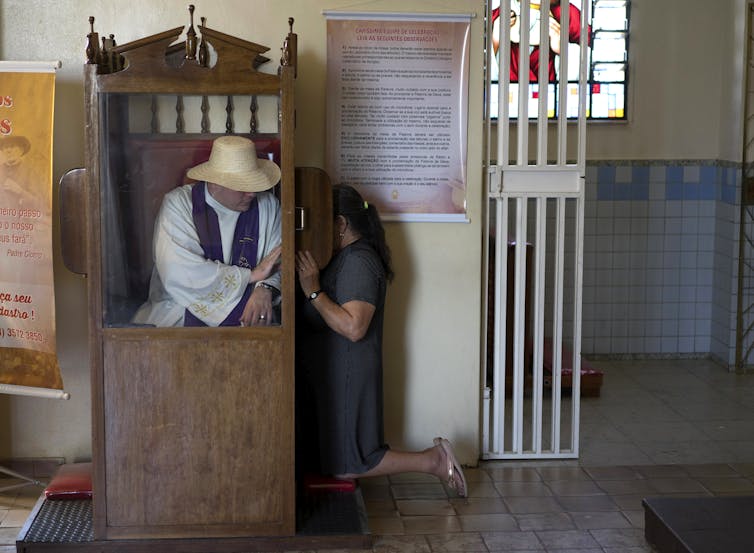 A man in a white robe and straw hat sits inside a wooden cubicle as a woman keels beside it, with a small screen shielding her face.