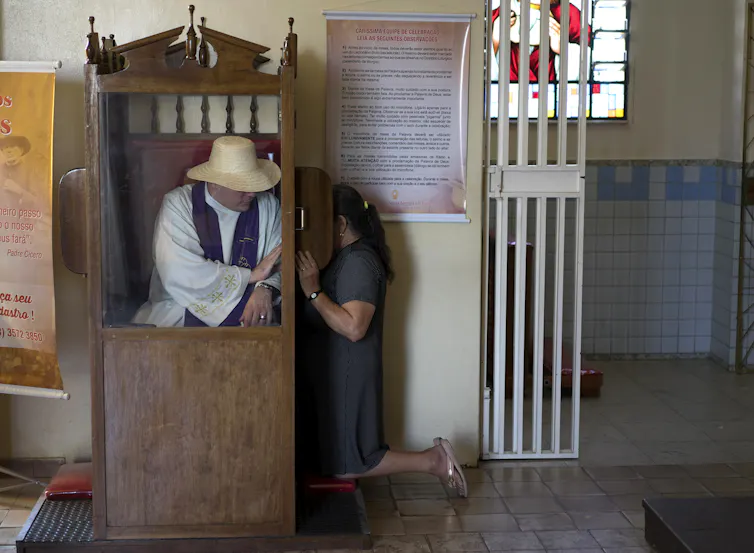 A man in a white robe and straw hat sits inside a wooden cubicle as a woman keels beside it, with a small screen shielding her face.
