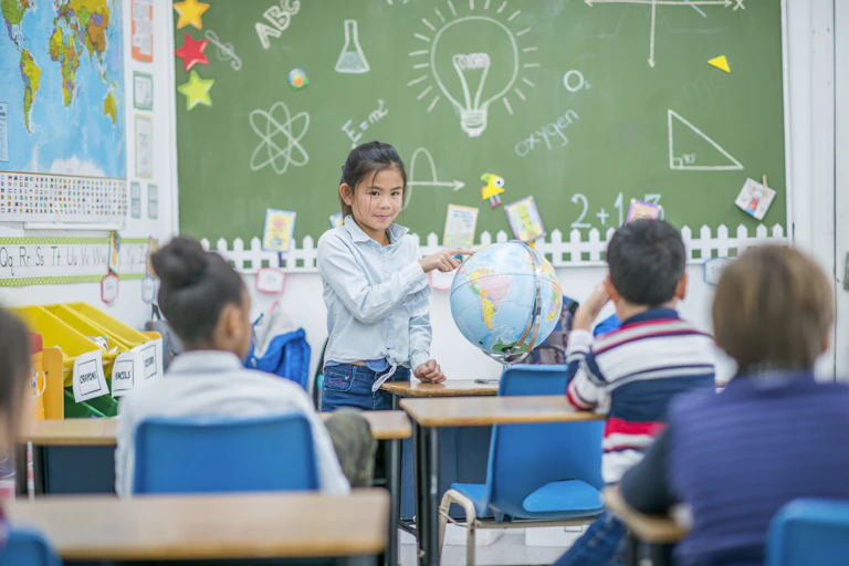 a student stands in front of other students sitting in a classroom and points to a location on a globe - a chalkboard with various drawings is behind her