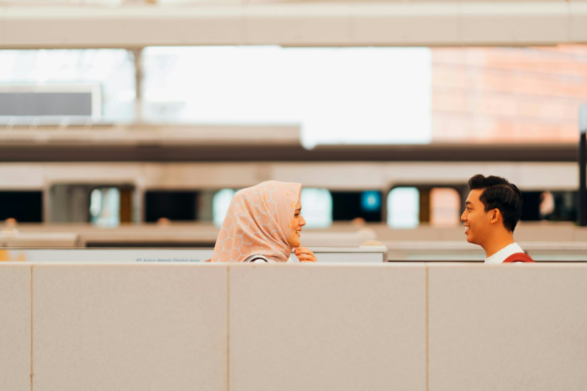 A woman in a hijab smiles at a man above a cubicle divider