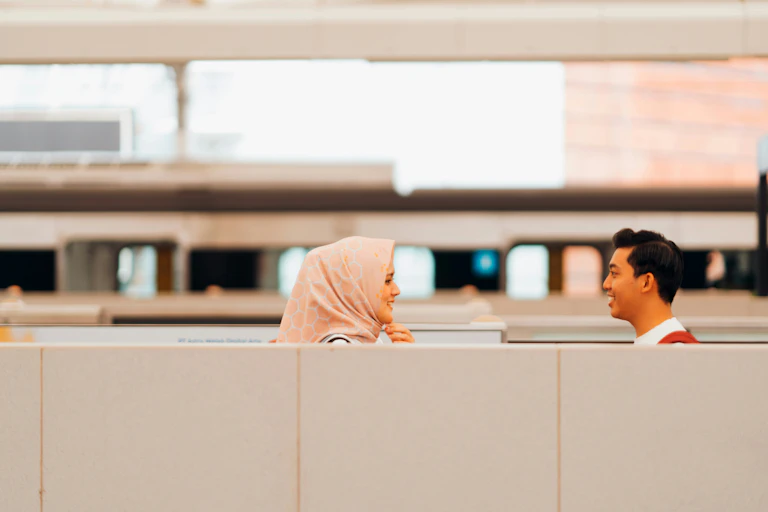 A woman in a hijab smiles at a man above a cubicle divider