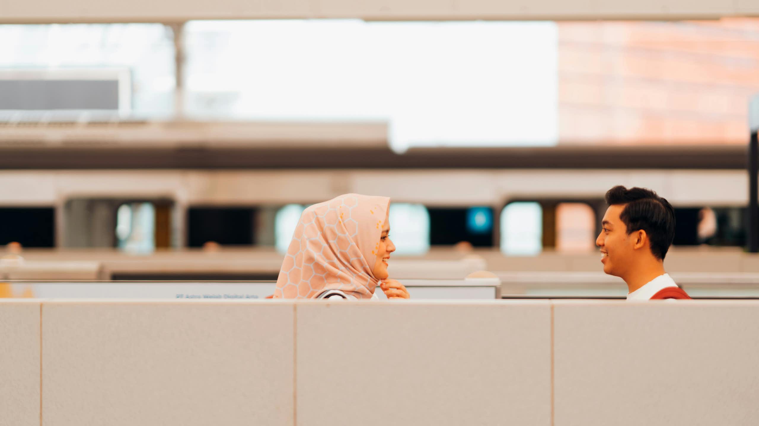 A woman in a hijab smiles at a man above a cubicle divider