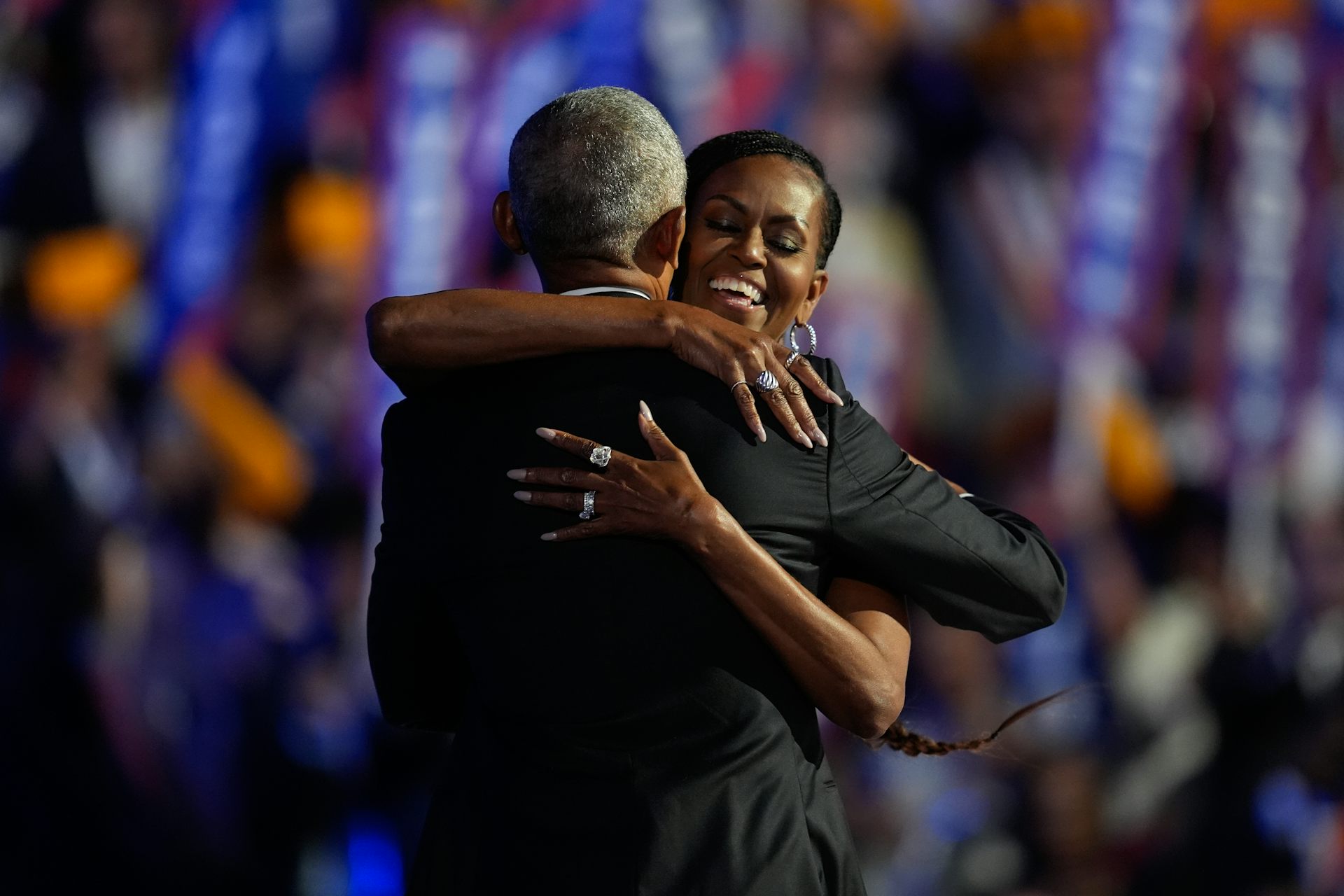 Mujer negra sonriente abraza al hombre negro, visto desde atrás y usa un traje, en el escenario frente a la multitud