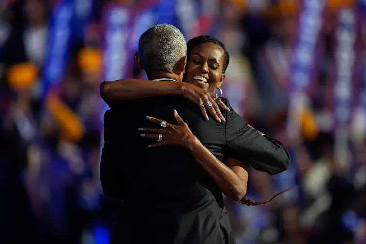 A smiling Black woman hugs a Black man, seen from behind and wearing a suit, on a stage in front of a crowd