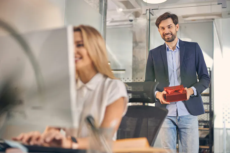 A man carrying a gift box walks up to a woman who is working at a computer