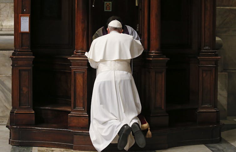 A man in white robes and a white skullcap, seen from the back, kneeling in front of an ornate wooden cubicle.