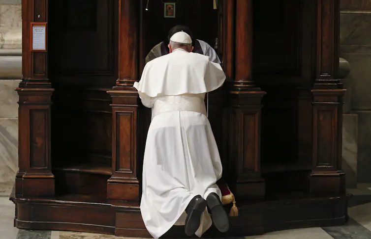 A man in white robes and a white skullcap, seen from the back, kneeling in front of an ornate wooden cubicle.