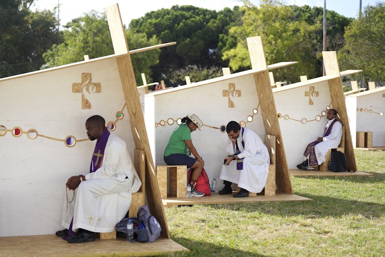 A row of open-air structures, with three walls for privacy, set up in a row, with a man in white robes sitting in each one.
