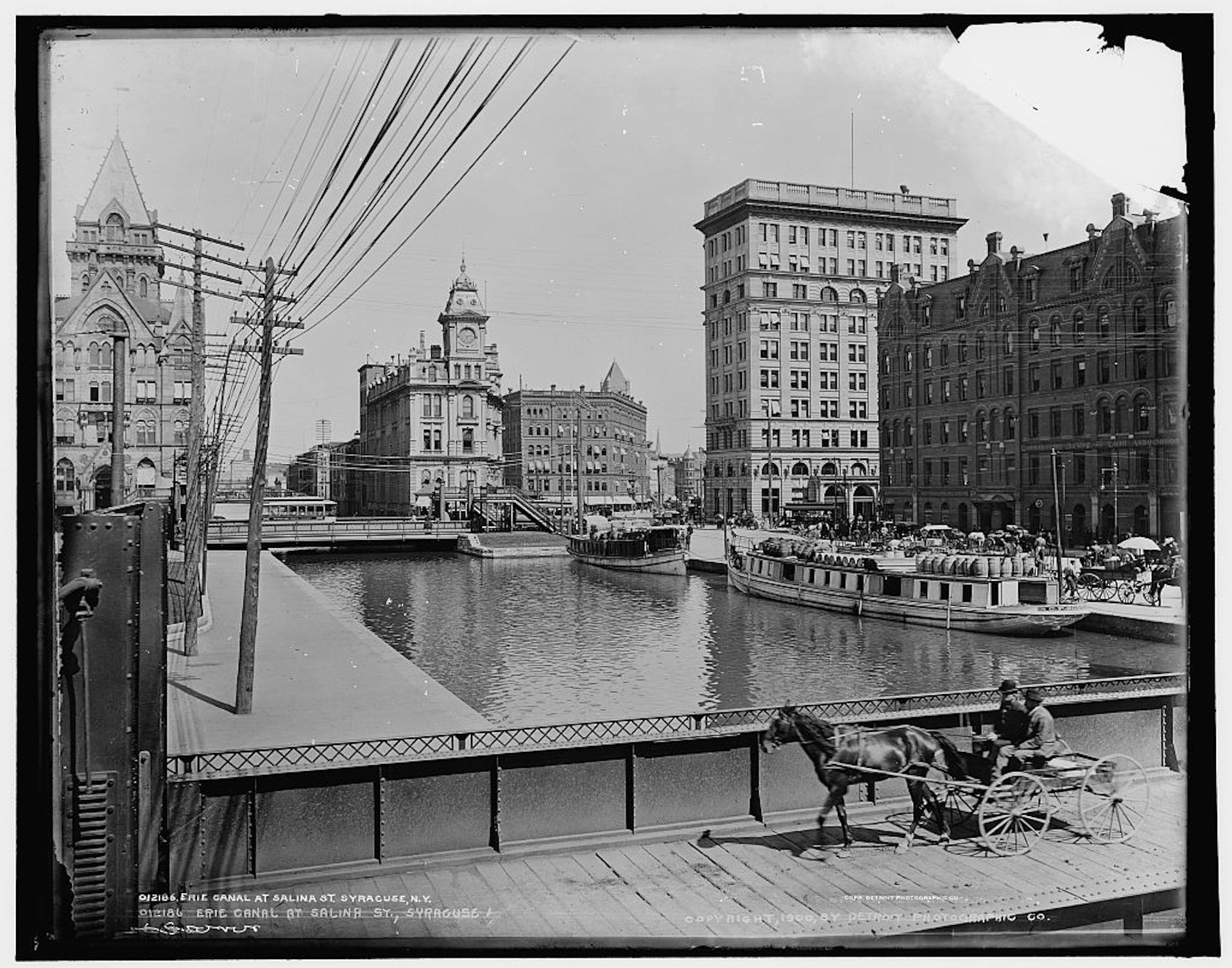 A person in a horse-drawn buggy rides over a bridge crossing the Erie Canal in downtown Syracuse. Barges are moored along the edge.
