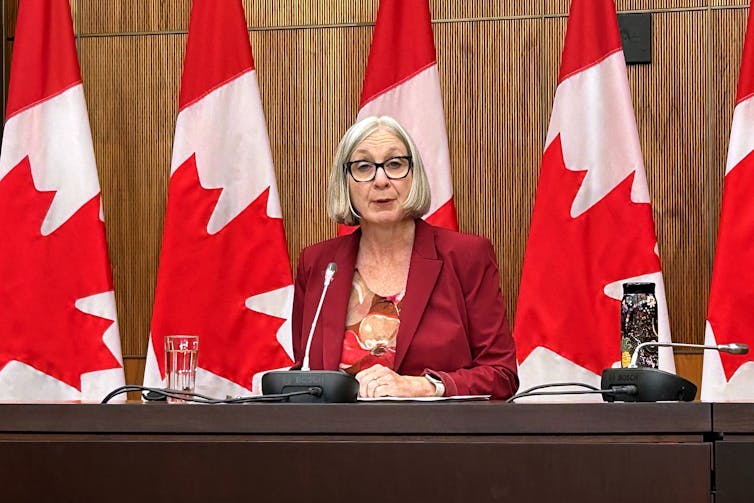La mujer blanca de mediana edad en el traje rojo dice mientras se sienta frente a la fila de banderas canadienses