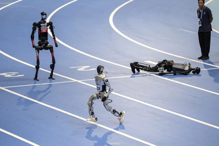 Three humanoid robots are on a race track, one standing, one going sideways and one flat on its face.