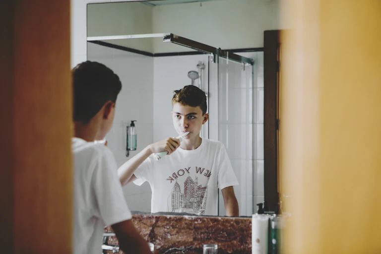 Teen boy brushes teeth looking bathroom mirror.