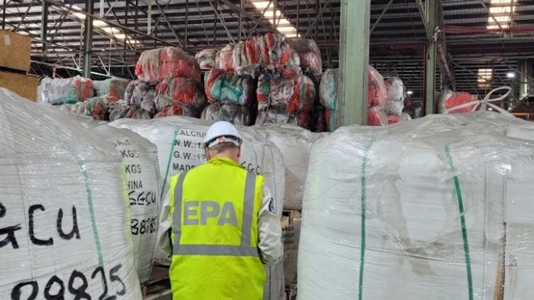 A man in a high-ves vest saying EPA, standing surrounded by giant bags of unrecycled plastic.