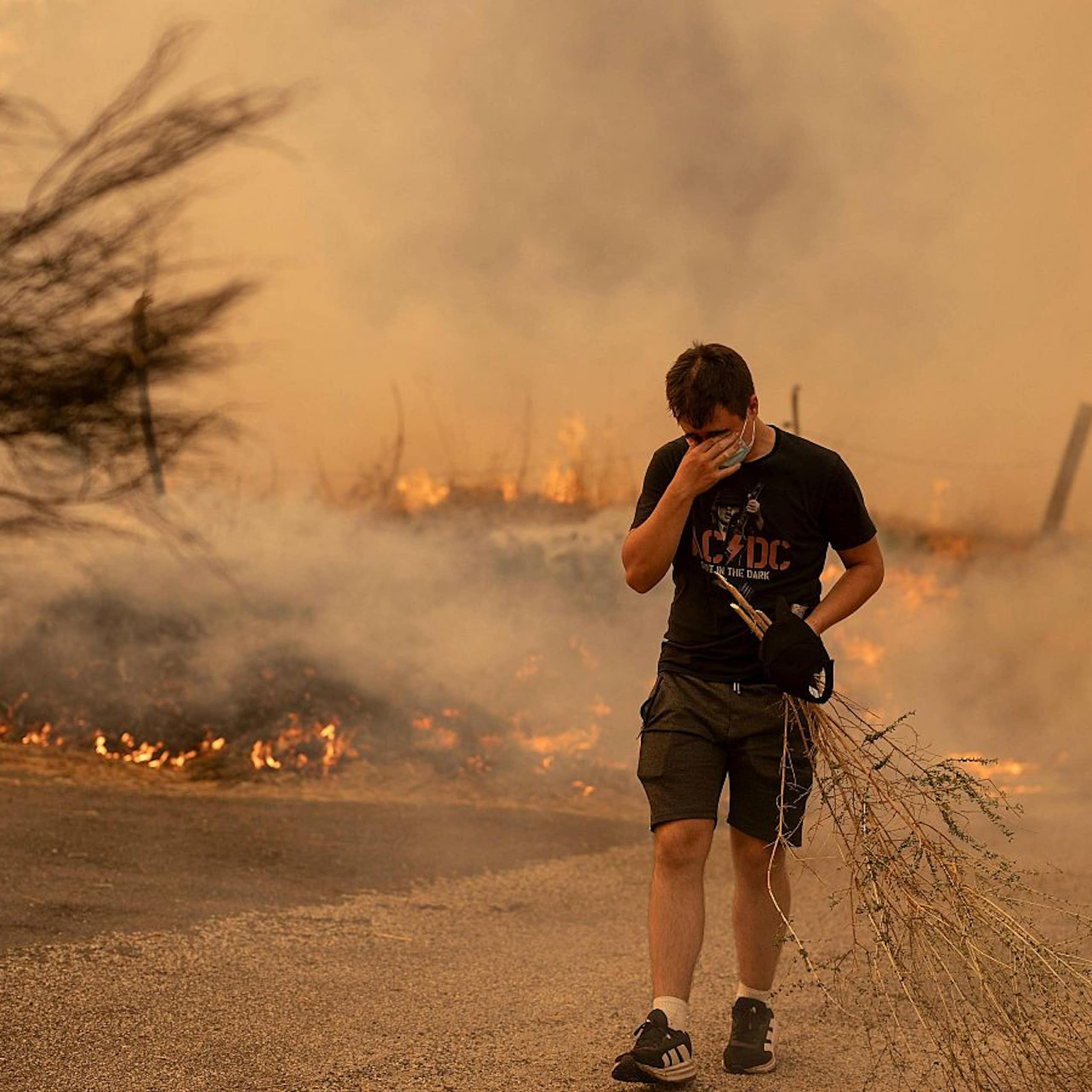 Un joven con mascarilla y una rama de arbusto camina en primer plano con llamas y humo de fondo