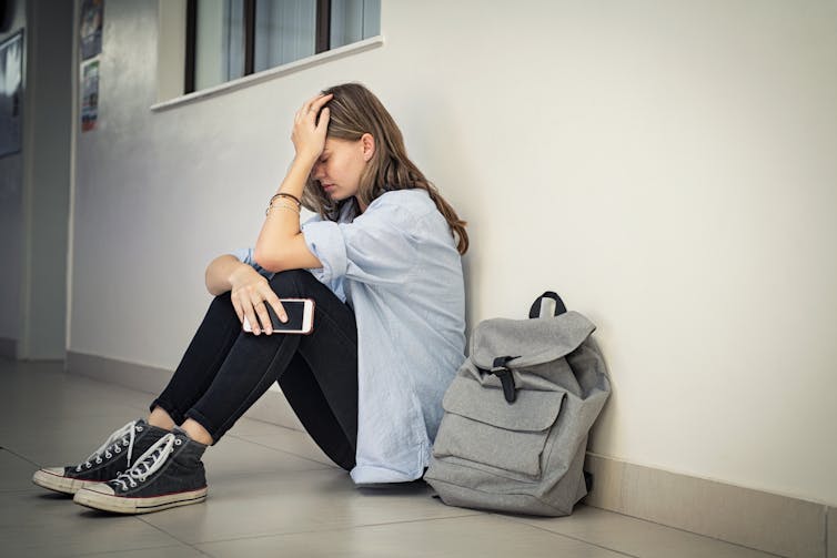 Upset and depressed girl holding smartphone sitting on college campus floor holding head.