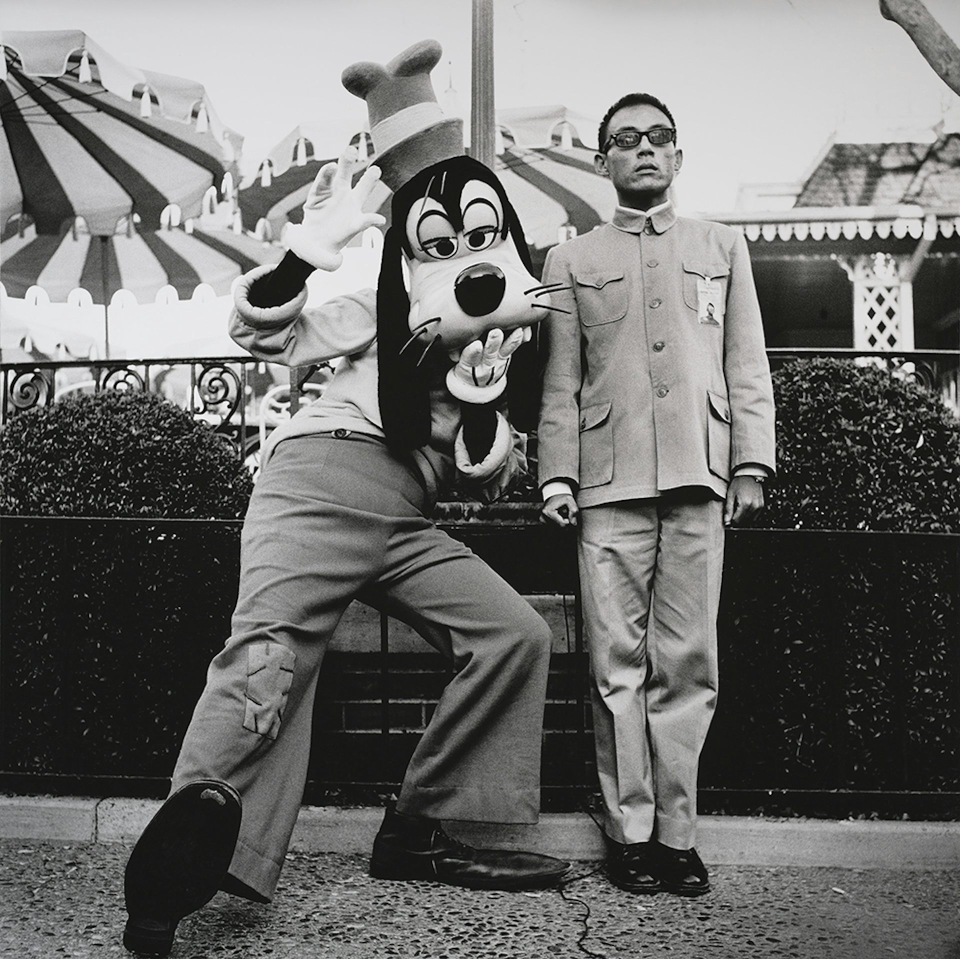 Young Asian man standing upright with serious expression on his face, posing next to a mascot of a Disney character making a silly pose.