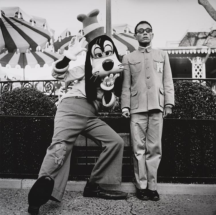 A immediately face, with a wink – the delicate humor of deadpan pictures 1 Young Asian man standing upright with serious expression on his face, posing next to a mascot of a Disney character making a silly pose.