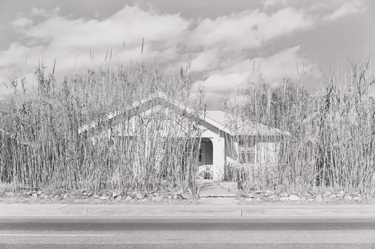 Black and white photograph of a stucco, single-story house surrounded by tall weeds.