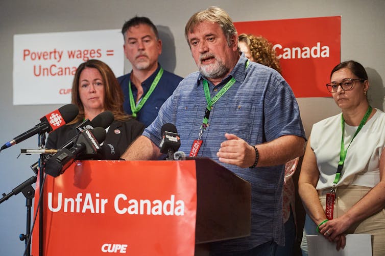 El hombre blanco con barba, que lleva la cinta, habló detrás del podio que dice que el injusto Canadá está en ella. Un grupo de personas también usa tiras detrás de él