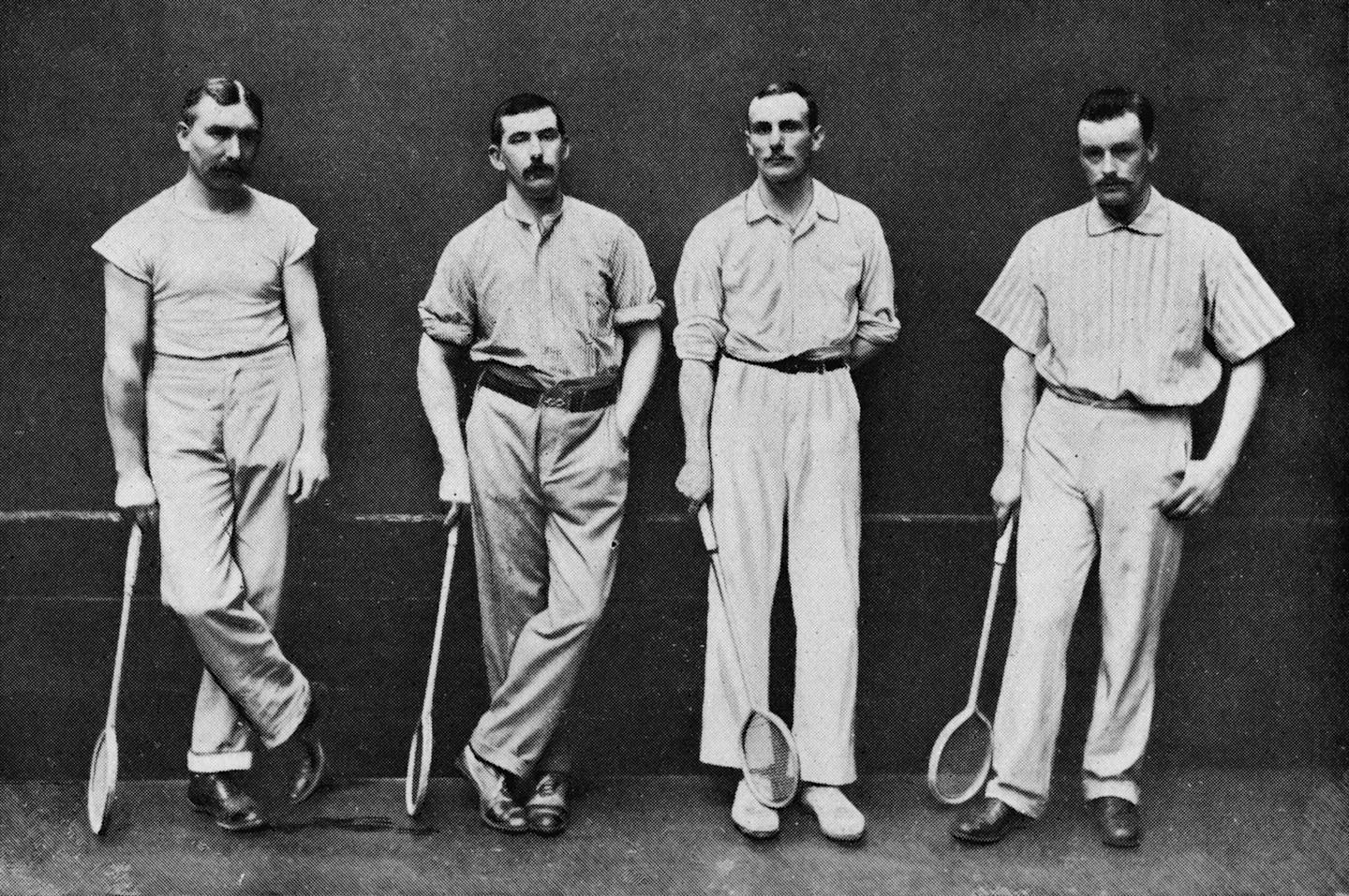 Black and white photo of four men in shirts, slacks and shoes holding squash rackets