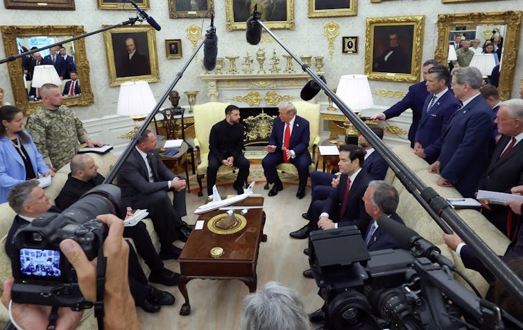 Microphone booms and cameras frame two men sitting on chairs.