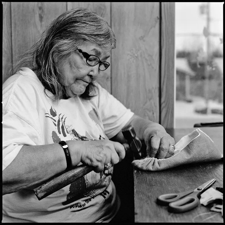 a black and white photo of an older woman working on a shoe at a table.