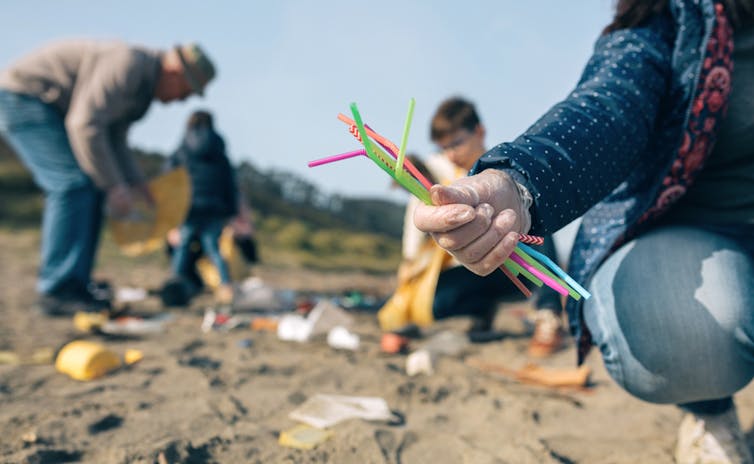 People cleaning litter on a beach. One holds a handful of plastic straws.