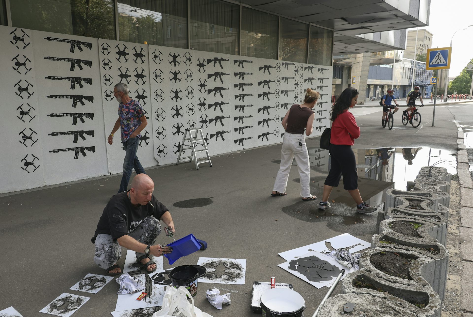 A man kneels in the street, making artworks out of old rifles, as passers-by look on.