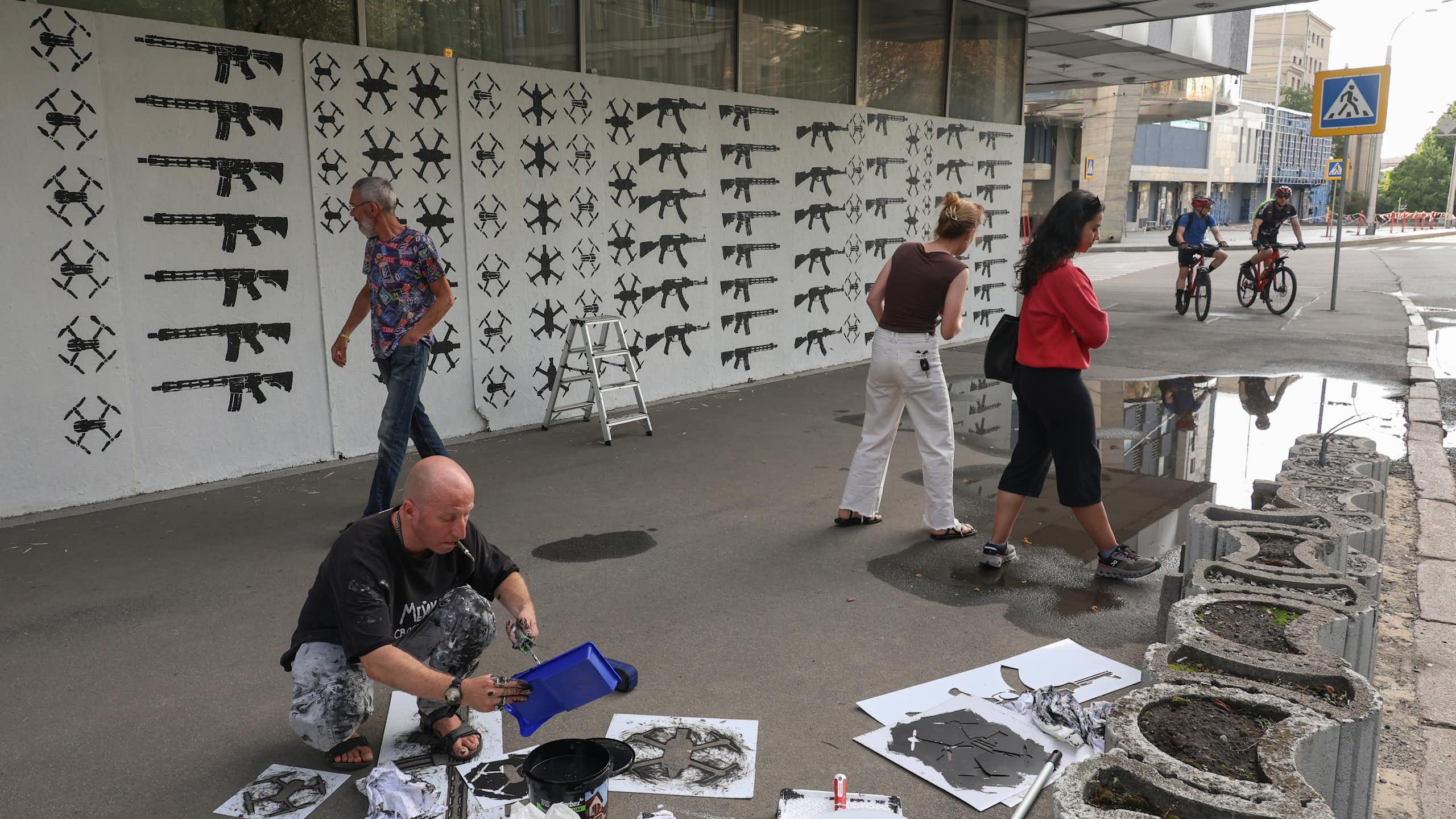 A man kneels in the street, making artworks out of old rifles, as passers-by look on.