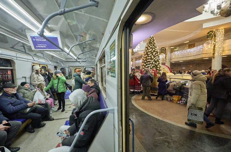 Men and women sit on an underground train in the Kharkiv metro. The metro station is decorated for Christmas.