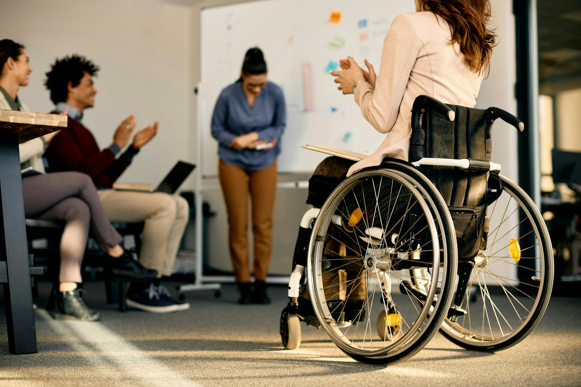 Une personne se tient devant un tableau blanc et s’incline tandis que d’autres, dont quelqu’un assis en fauteuil roulant, l’applaudissent