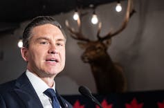 Un hombre de cabello oscuro le habla a una cabeza de ciervo disecada en la pared detrás de él.