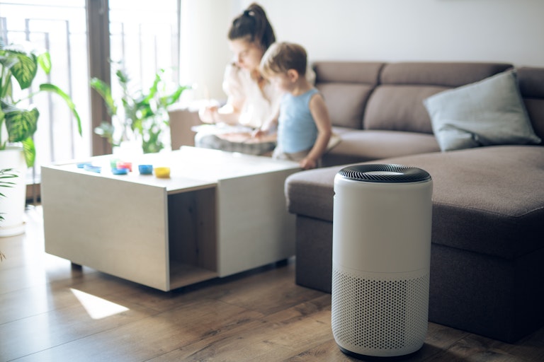 An air purifier sits in the foreground while a mother and child spend time together on a couch in the background