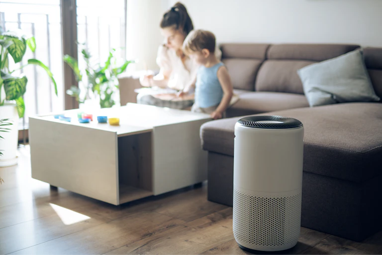 An air purifier sits in the foreground while a mother and child spend time together on a couch in the background