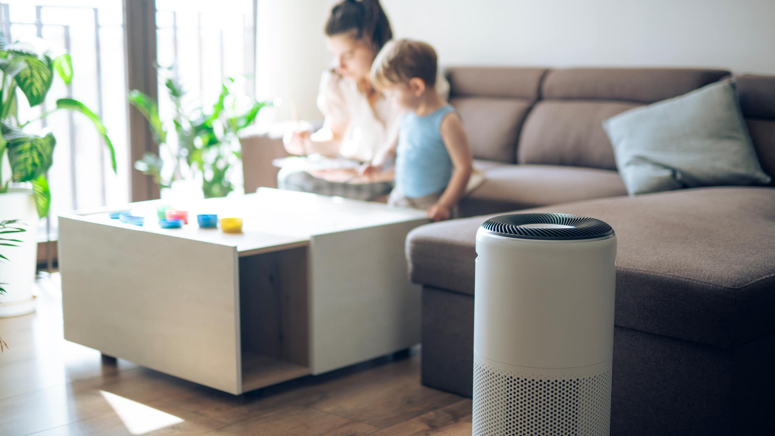 An air purifier sits in the foreground while a mother and child spend time together on a couch in the background