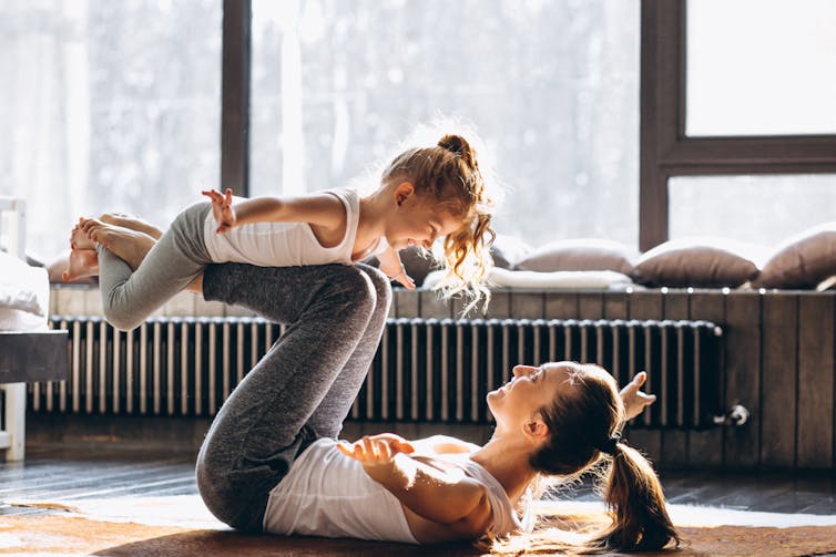 A woman lies on the floor, balancing her daughter on her shins.