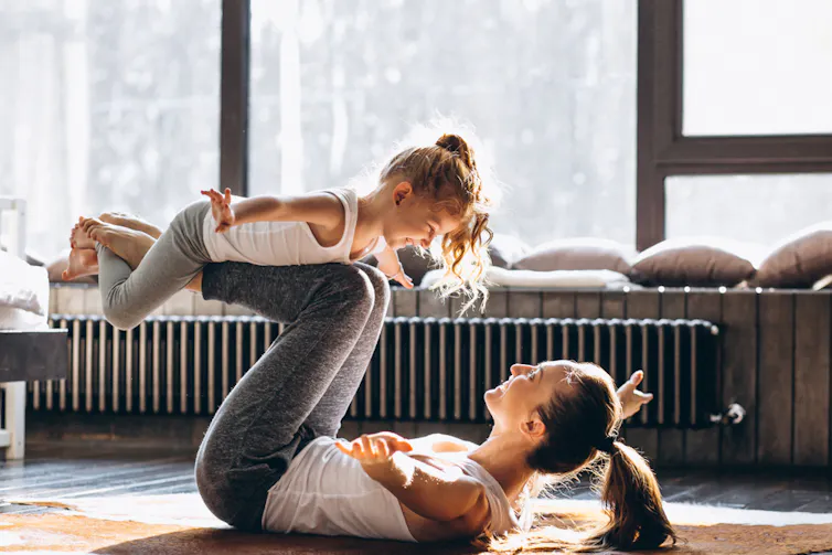 A woman lies on the floor, balancing her daughter on her shins.