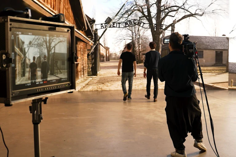 A camera monitor set up behind two people walking in front of a large screen showing the entrance to Auschwitz