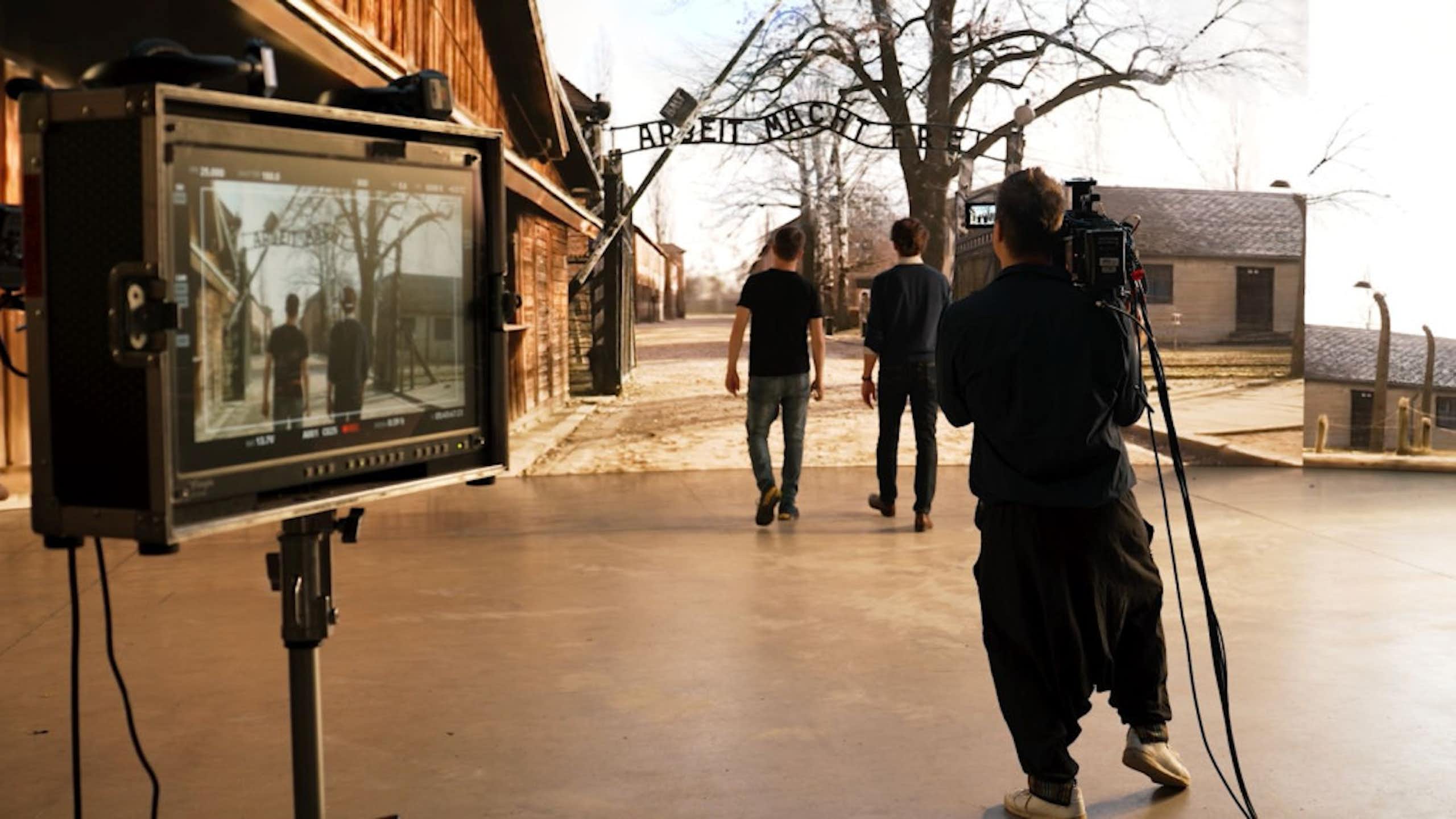 A camera monitor set up behind two people walking in front of a large screen showing the entrance to Auschwitz