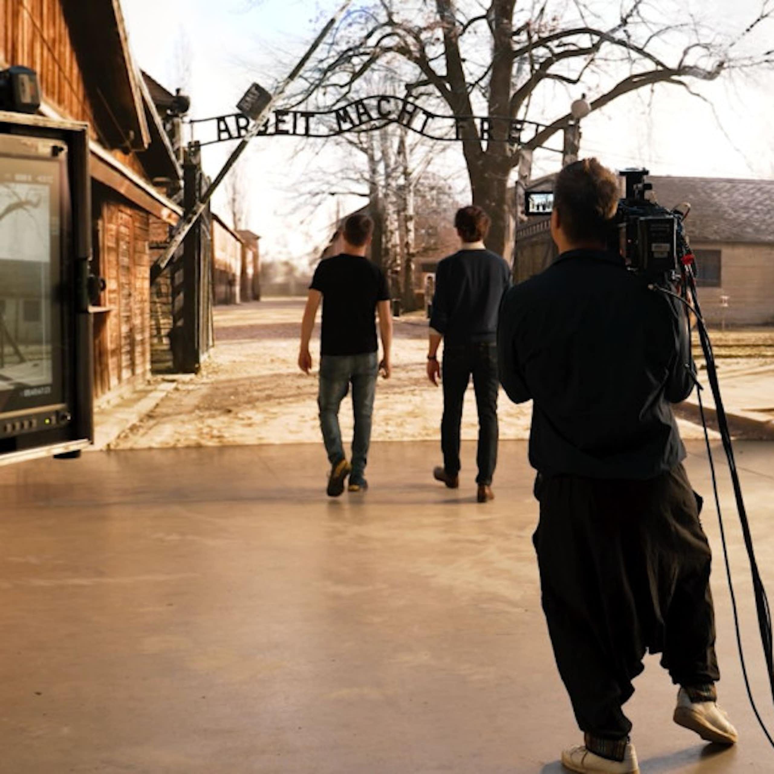 A camera monitor set up behind two people walking in front of a large screen showing the entrance to Auschwitz