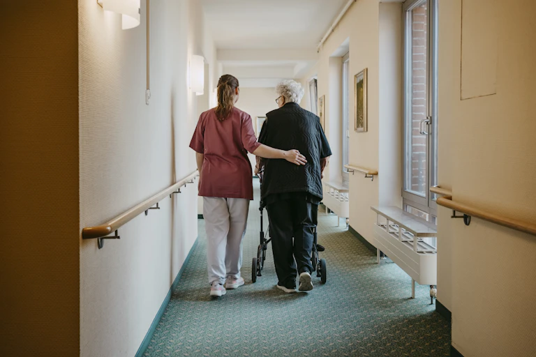 Un aide-soignant accompagne une femme âgée équipée d'un déambulateur dans un couloir.